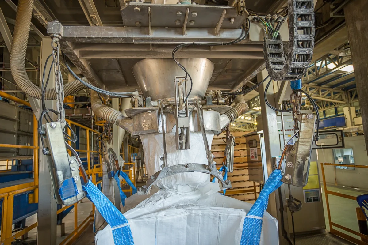 A bulk bag receiving chemical powder beneath industrial discharge equipment.